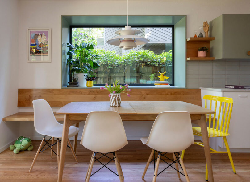 Comedor moderno con una mesa de madera, sillas blancas y amarillas, una vista de ventana de vegetación y plantas en el alféizar. La luz colgante cuelga arriba.