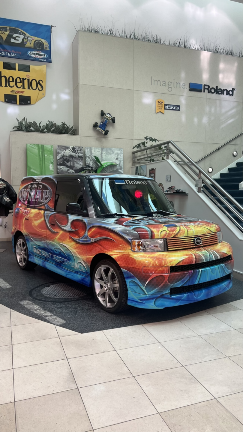 A colorful, graffiti-style painted car displayed indoors near stairs, with posters and greenery in the background