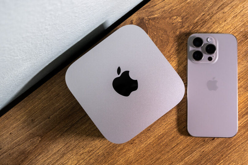 A silver Apple Mac Mini and a silver iPhone with triple camera lenses are placed on a wooden surface.