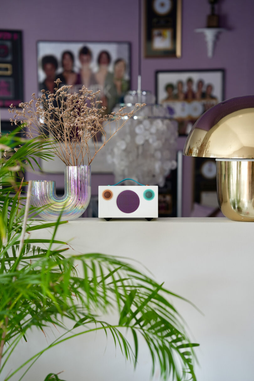 A modern living room with a purple wall, a table with a colorful vase, a small speaker, and a gold lamp. Green plants in the foreground add a touch of nature.