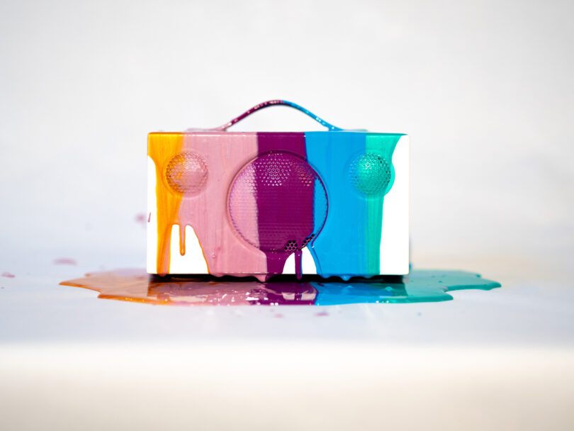 A white speaker covered in dripping paint of various colors including orange, purple, blue, and teal on a light background.