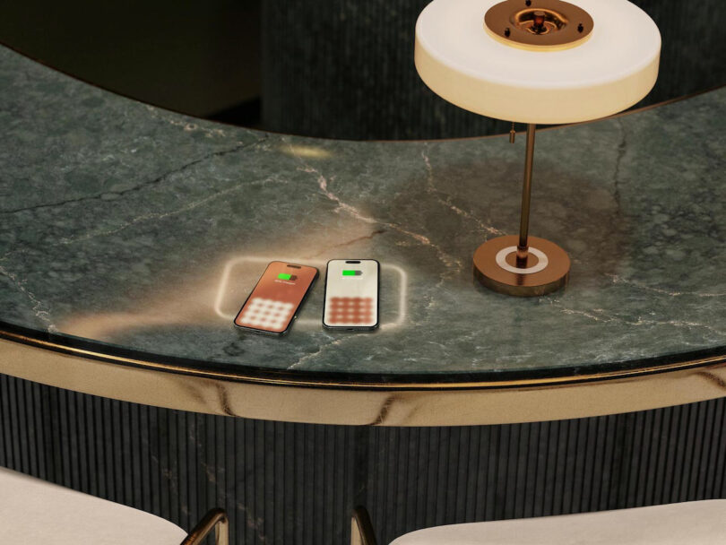 Two smartphones charging wirelessly on a marble surface beside a round lamp with a white shade.