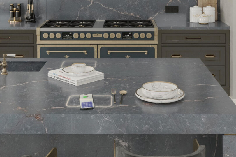 A modern kitchen with a dark marble island, cookbook, phone, and table setting. A dual-oven stove with metallic accents is in the background.