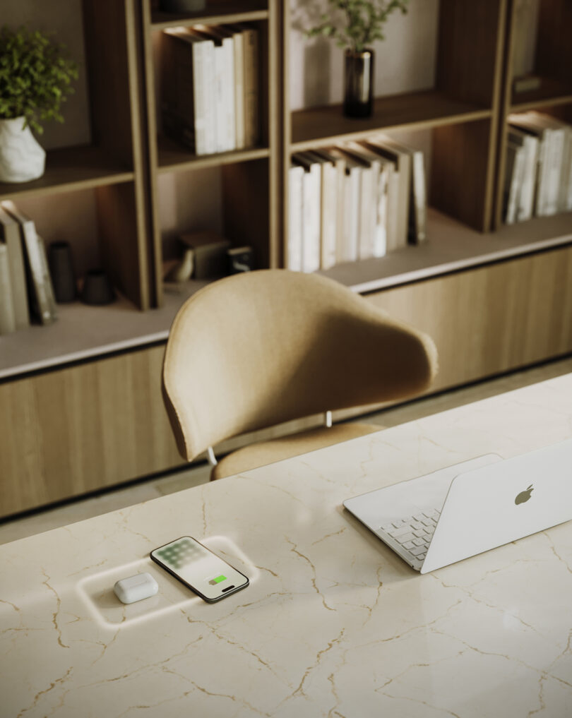 A marble desk with a white laptop, smartphone, and wireless earbuds. A wooden chair is tucked in, and a bookshelf with decorative items and books is in the background.