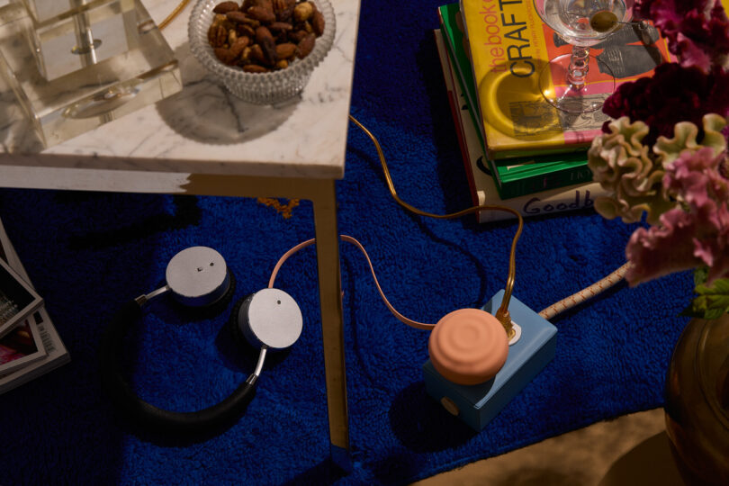 A living room scene with a table holding a bowl of nuts, headphones, books, flowers, and a blue and peach electronic device on a blue carpet.