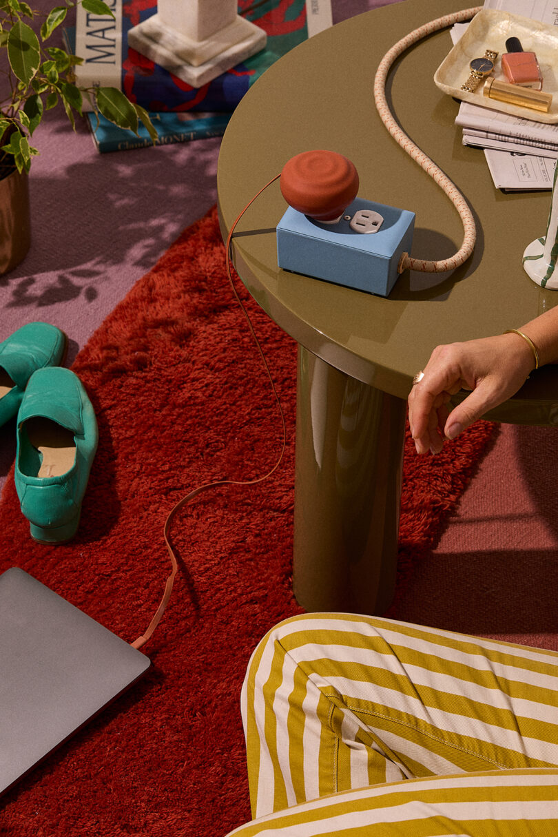 A person sits on a red rug, wearing striped pants. A table holds a power adapter, papers, and a drink. Nearby are green slippers and a laptop.