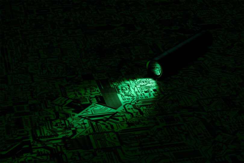 A flashlight shines on a dark, intricate circuit board, highlighting its patterns.