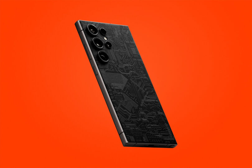 Black smartphone with a circuit board design on the back, featuring multiple rear cameras, displayed diagonally against a solid orange background.