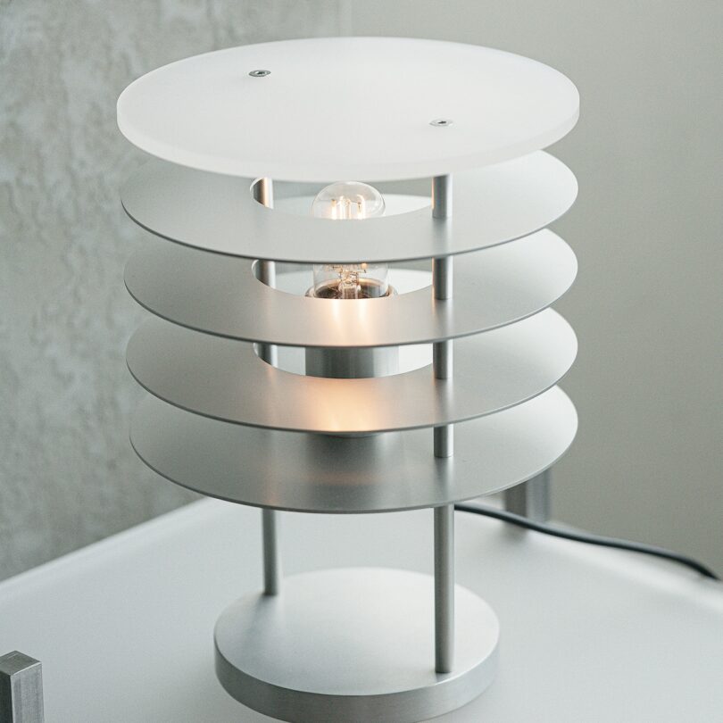 A modern, round table lamp with a layered metallic design and a visible light bulb, placed on a light-colored surface.