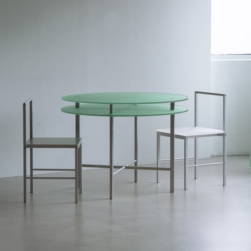 A round, translucent green table with two metal-framed chairs, one with a green seat and the other white, in a minimalist room with a window.