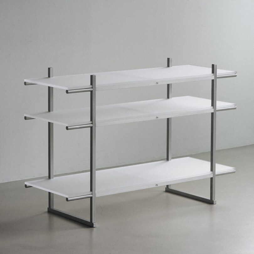 A minimalist, three-tiered shelving unit with a metal frame and white shelves, standing in a neutral-toned room.