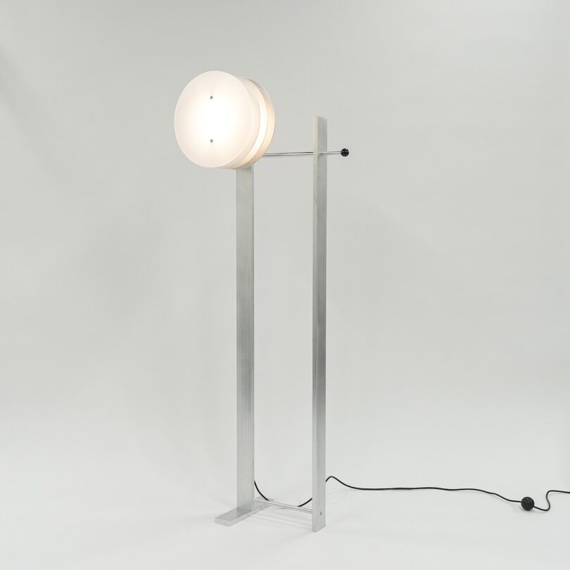 A modern freestanding lamp with a circular, frosted glass shade and a minimalist metal frame on a plain background. The lamp's cord is visible on the floor.