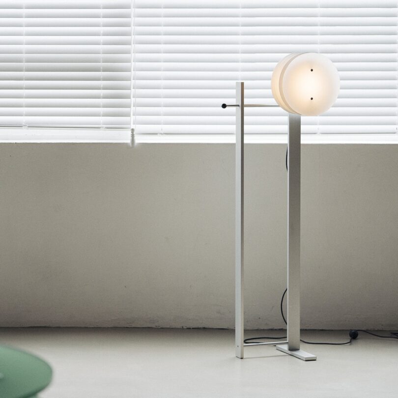 Minimalist floor lamp with a round white shade beside a wall with closed white blinds.