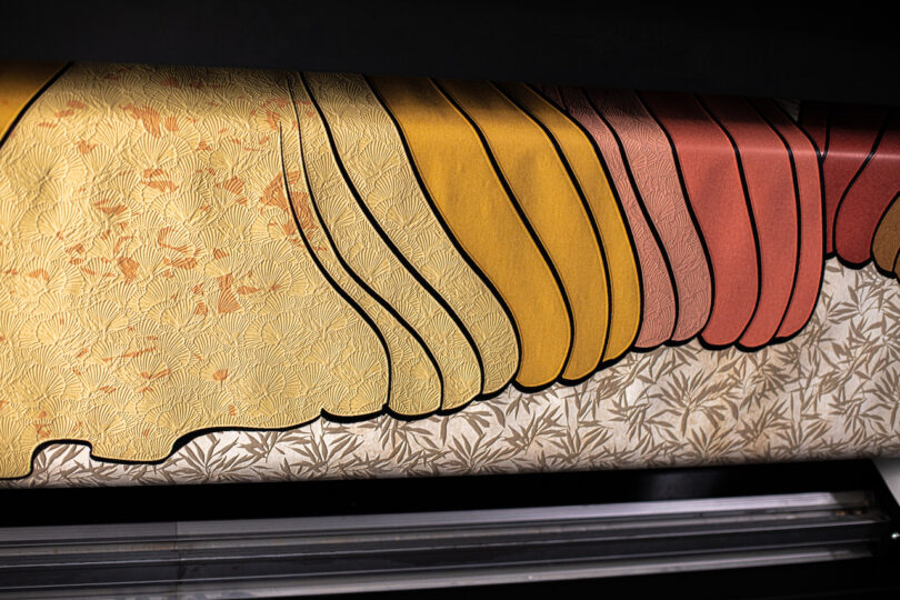 Close-up of a textile printing machine producing fabric with layered, scalloped patterns in shades of yellow, orange, and pink, featuring intricate leaf designs