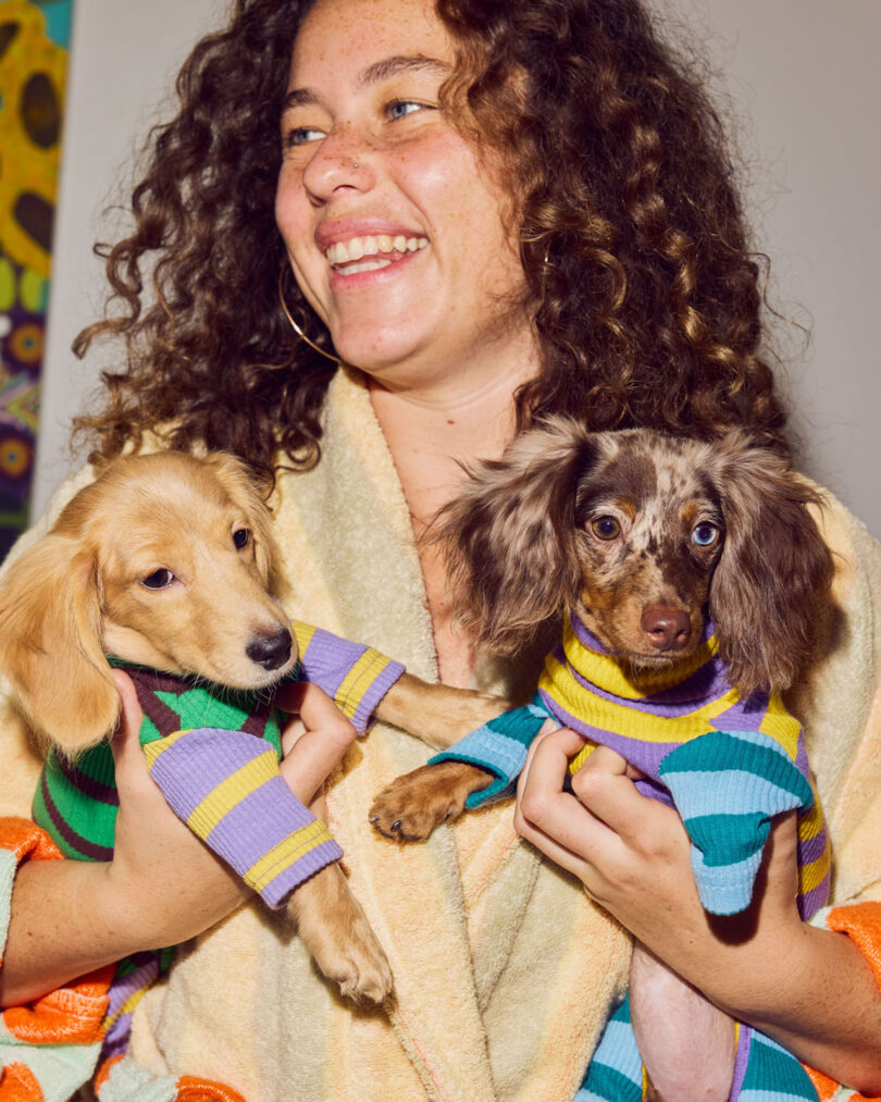 A woman smiles while holding two dachshund puppies in striped sweaters