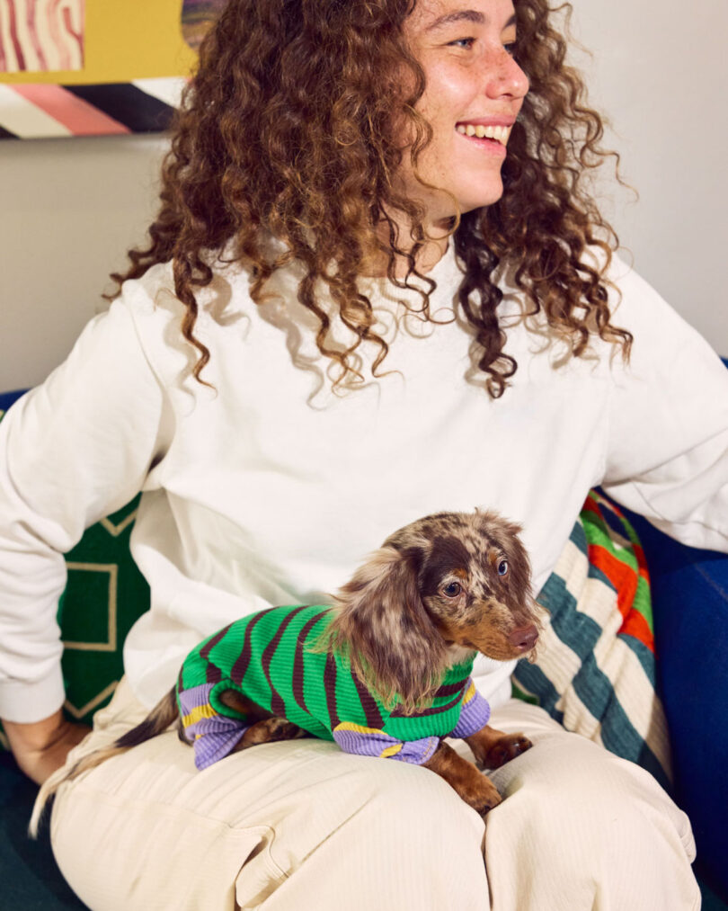 A person sits on a couch, smiling, with a small dog wearing a green and purple striped sweater on their lap