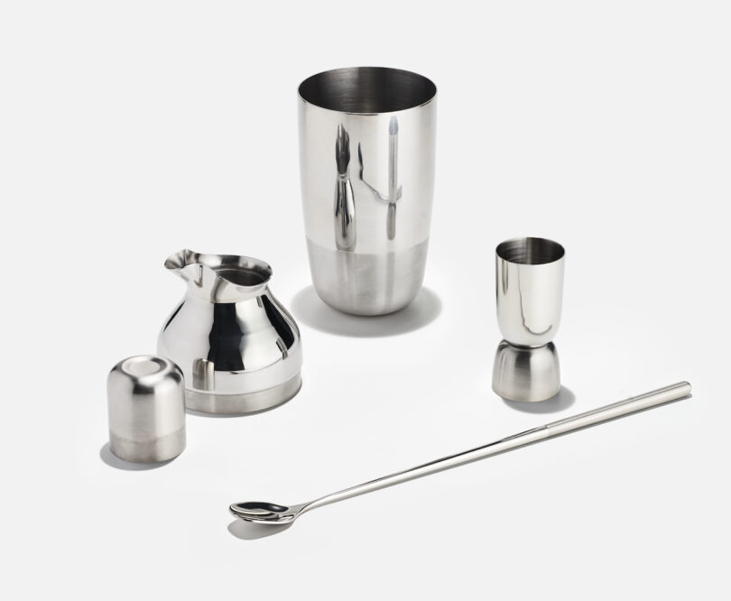 A set of stainless steel bar tools, including a shaker, jigger, strainer, capped container, and stirring spoon, arranged on a white background