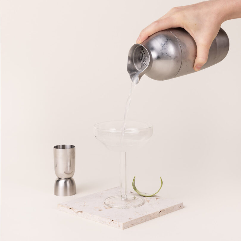 A hand pouring liquid from a metal cocktail shaker into an empty glass on a stone coaster. A metal jigger and lemon peel are beside the glass