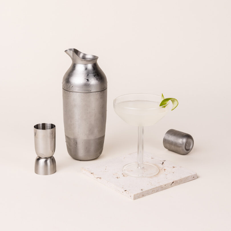 Stainless steel cocktail shaker, jigger, and strainer next to a cocktail glass with a lime garnish on a white surface