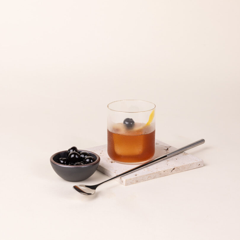 A glass of iced tea with a cherry garnish sits on a coaster, next to a small bowl of cherries and a metal spoon