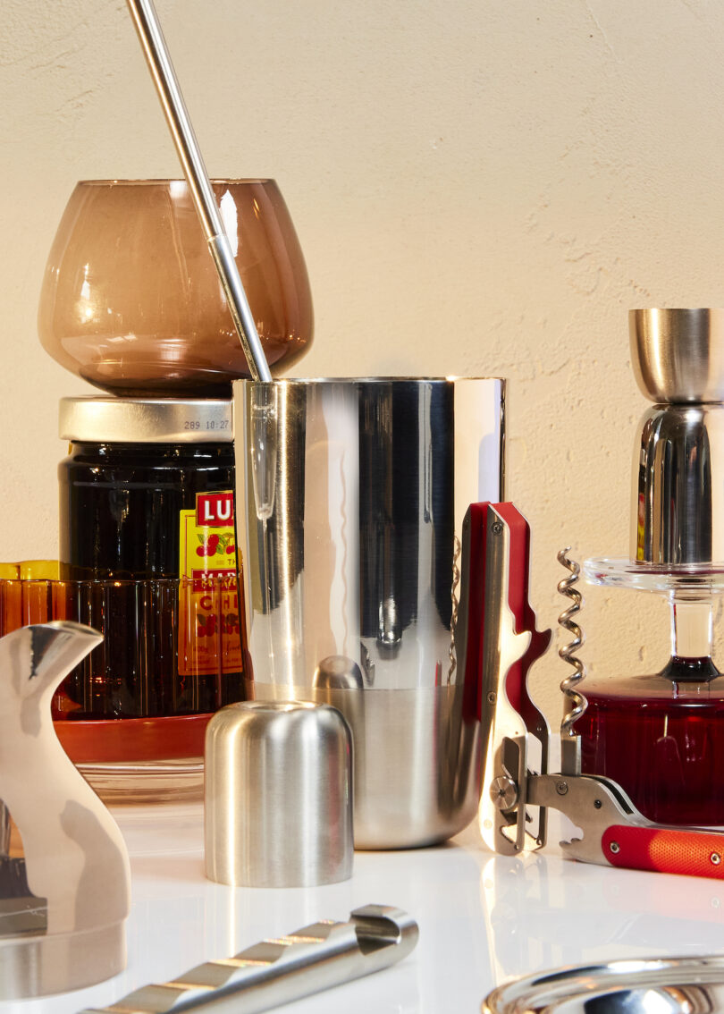 A collection of bar tools and glassware, including a cocktail shaker, a corkscrew, various bottles, and a jigger, arranged on a light surface