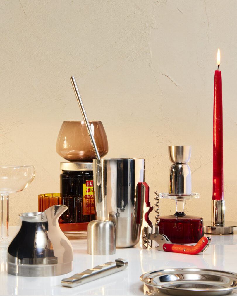 A collection of bar tools and accessories on a table, including a cocktail shaker, corkscrew, jigger, and a lit red candle