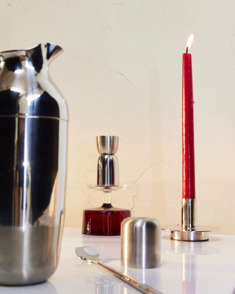 A cocktail shaker, jigger, red taper candle, and mixing spoon are arranged on a white surface against a beige wall