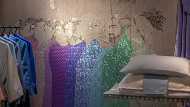 A wall mural depicting seven stylized figures in patterned clothing next to a clothes rack and folded blankets