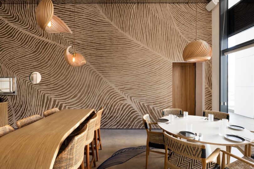 A modern dining area with a wooden table, wicker chairs, and a textured wall pattern. Hanging pendant lights add warmth to the space. Large window on the right