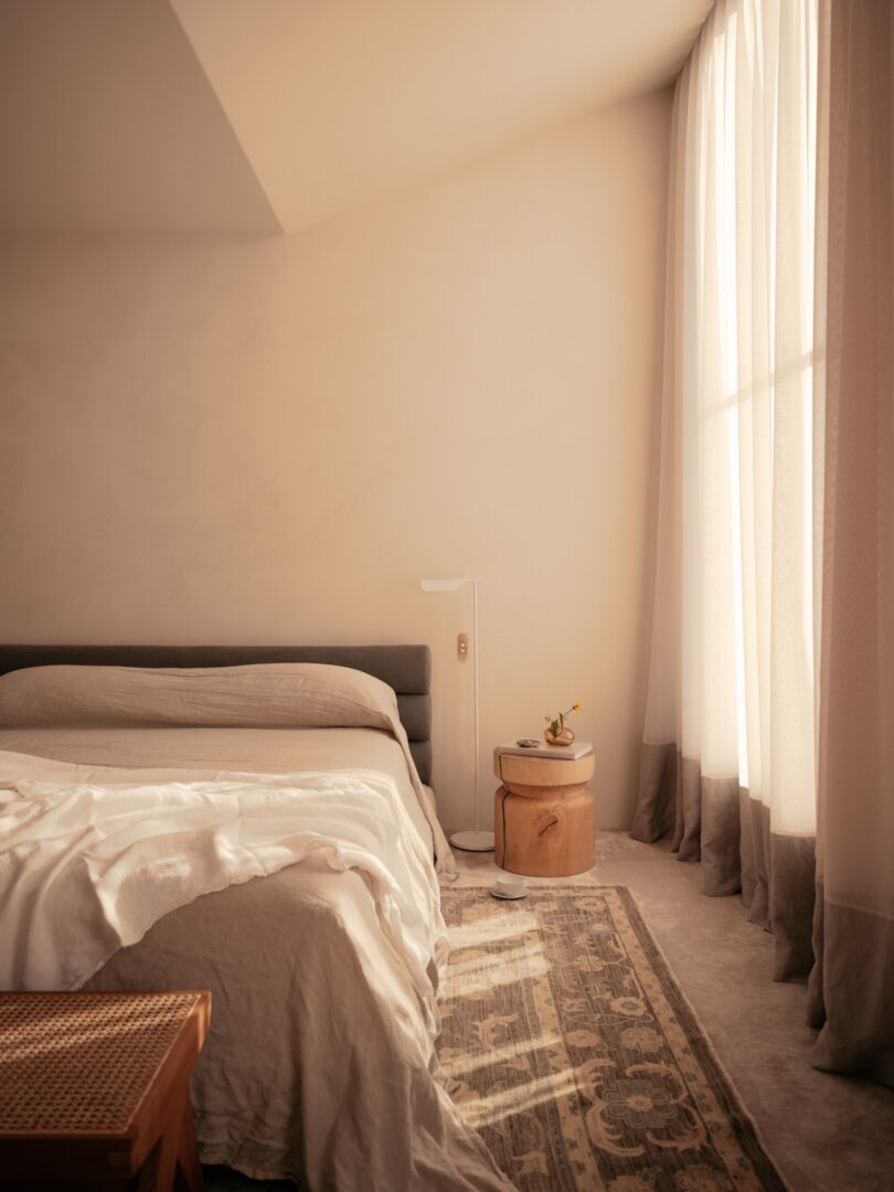 A softly lit bedroom featuring a neatly made bed, a wooden nightstand with a small lamp and plant, and sheer curtains letting in natural light.