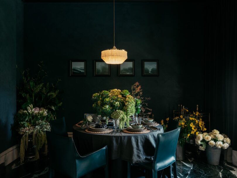 Dimly lit dining room with a round table set for a meal, surrounded by dark chairs. The table and surrounding area are decorated with various green and yellow plants.