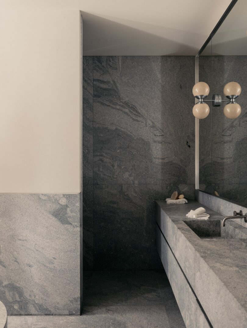 A modern bathroom with gray stone walls and countertops, featuring a large mirror, double sink, and spherical light fixtures.