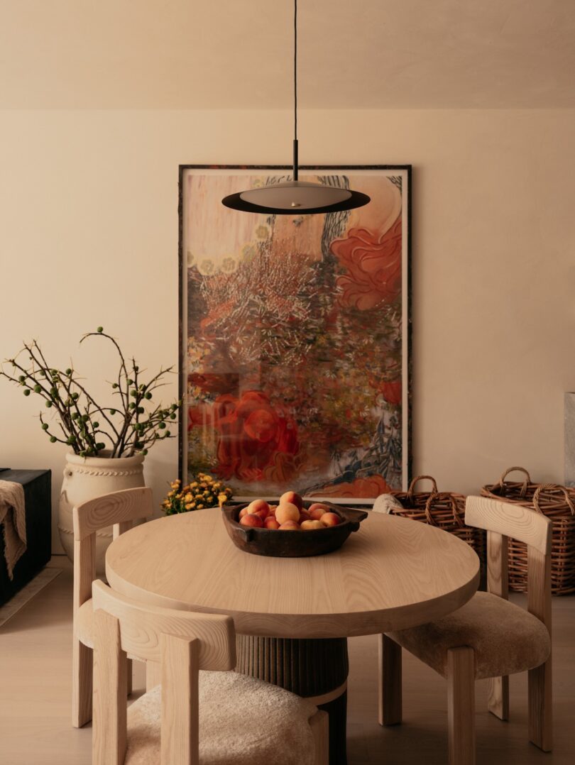 A dining area with a round wooden table, four chairs, and a bowl of fruit. A large, colorful painting hangs on the wall behind. A potted plant and woven baskets are in the background.