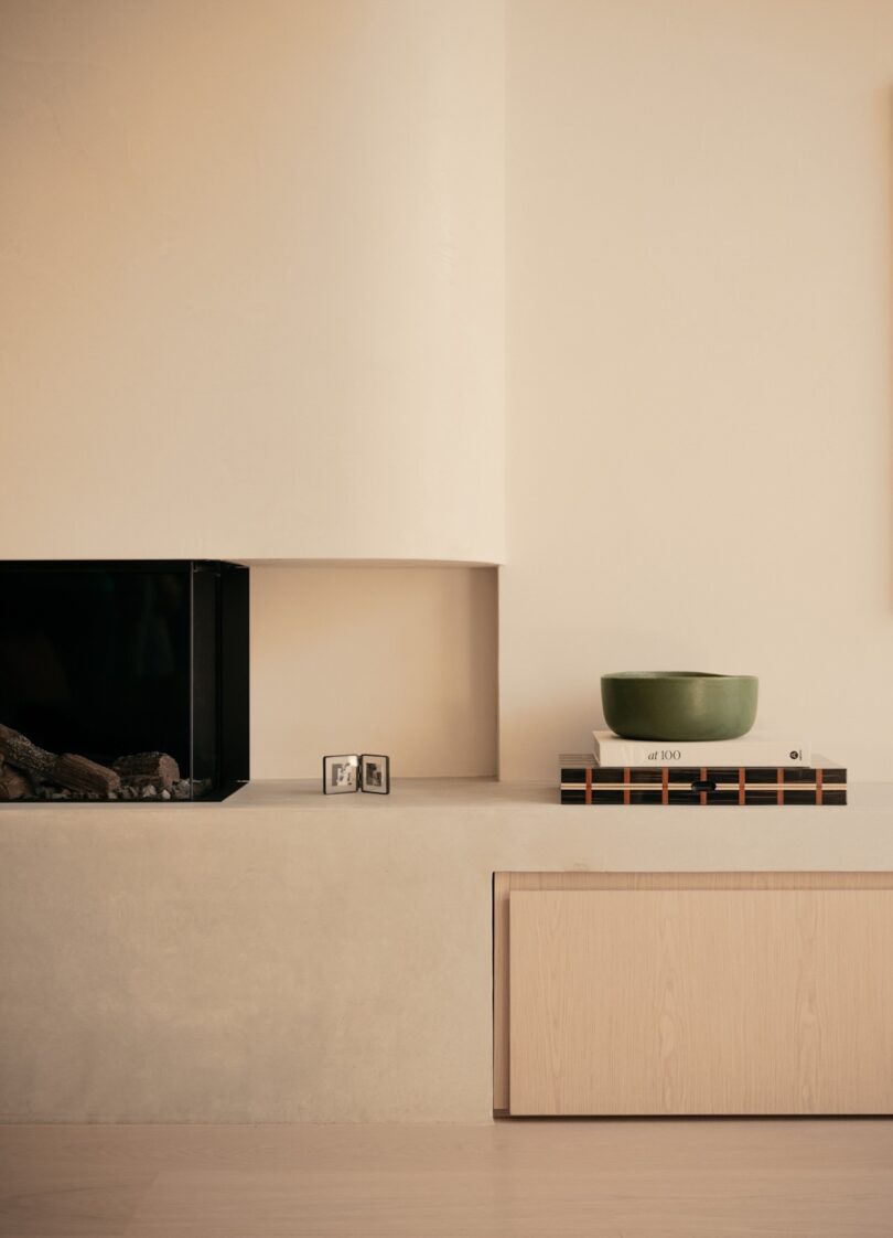 Minimalist living room with a fireplace, small framed picture, stacked books, and a green bowl on a beige shelf. Wooden cabinet below.