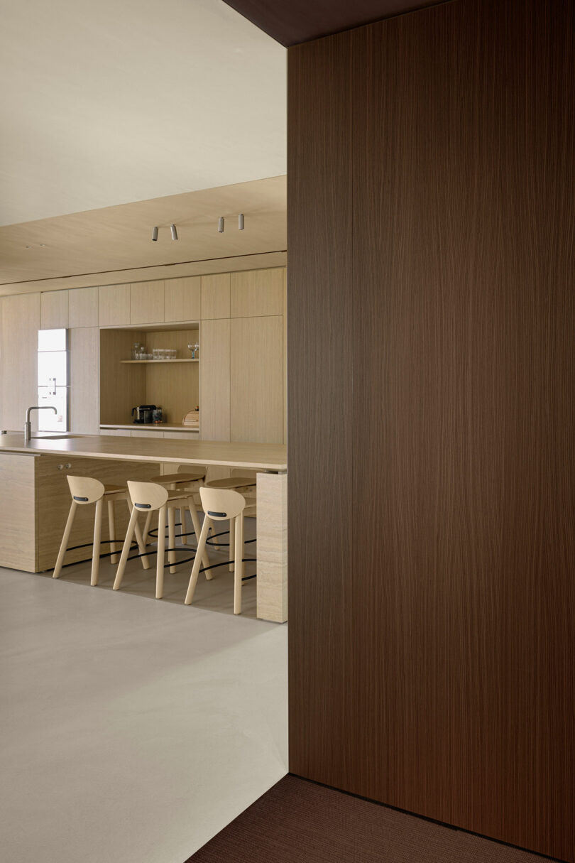 Minimalist kitchen with light wood cabinetry, a large island with three chairs, and modern fixtures. A wooden wall panel is in the foreground.