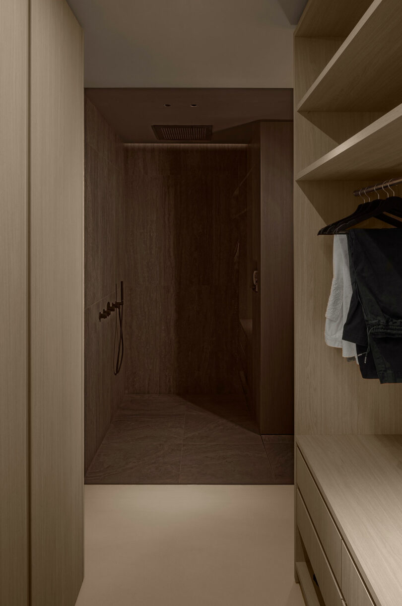 Minimalist wooden walk-in closet with hanging clothes, drawers, and an adjacent stone-tiled shower area.