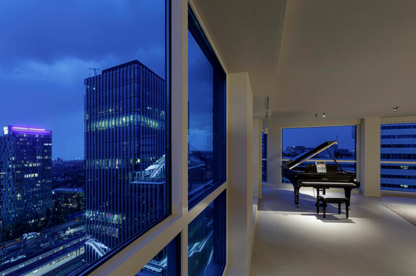 A grand piano is placed in a modern, spacious room with large windows offering a nighttime city view.