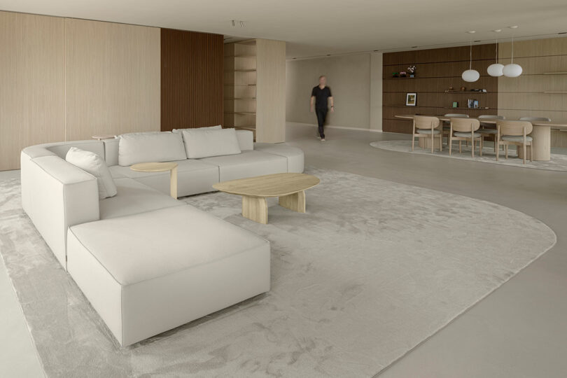 Modern living room with beige sofa, wooden coffee tables, and a dining area in the background. A person is walking across the room.