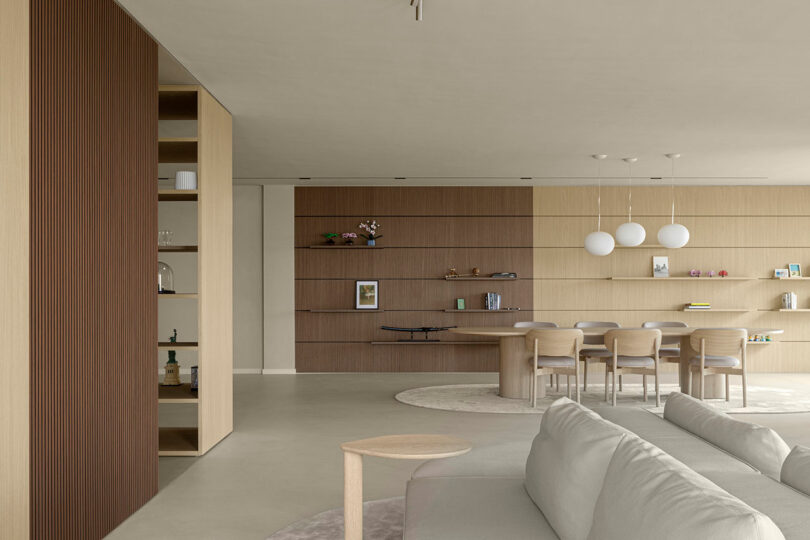 A modern living and dining area with beige and wooden tones, featuring a sofa, a dining table with chairs, and decorative shelving with books and items. Three spherical pendant lights hang above.