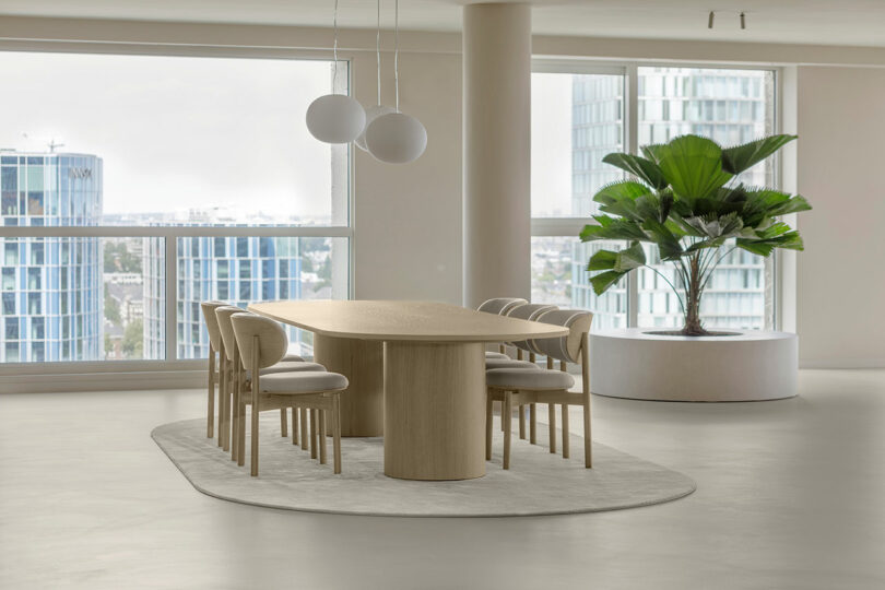 A modern dining area with a wooden table and six chairs on a light rug. Two ceiling lights hang above, and a potted plant is near the windows overlooking a cityscape.