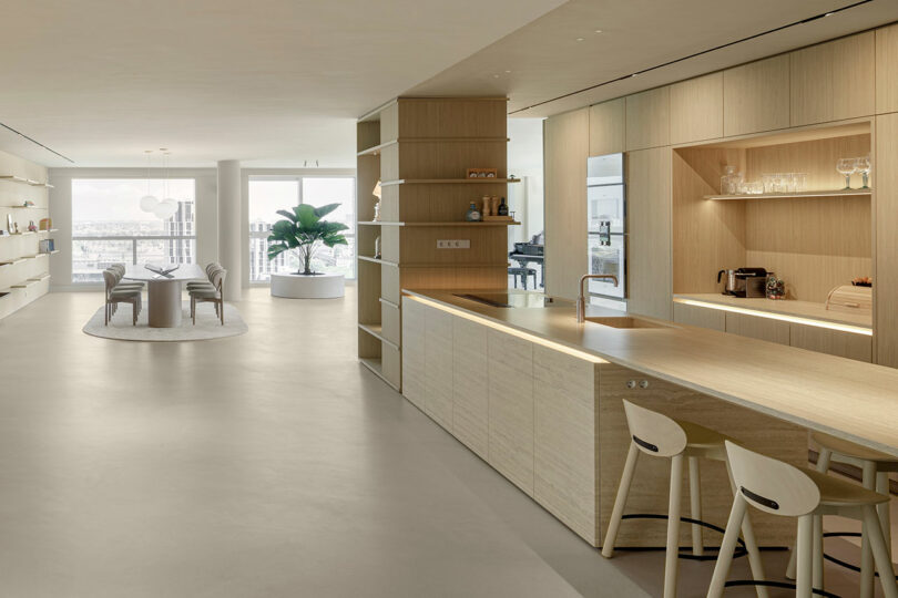 Modern open-plan living space with a minimalist kitchen and dining area. Light wood finishes, built-in shelves, and a city view through large windows enhance the airy ambiance.