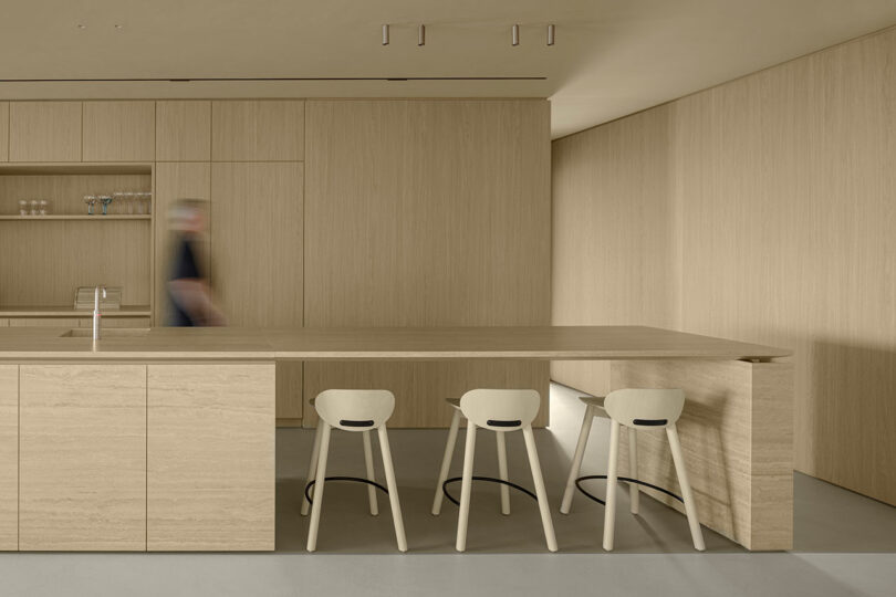 Minimalist kitchen with light wood cabinets and a large matching island. Three bar stools are at the island, and a blurred figure walks by.