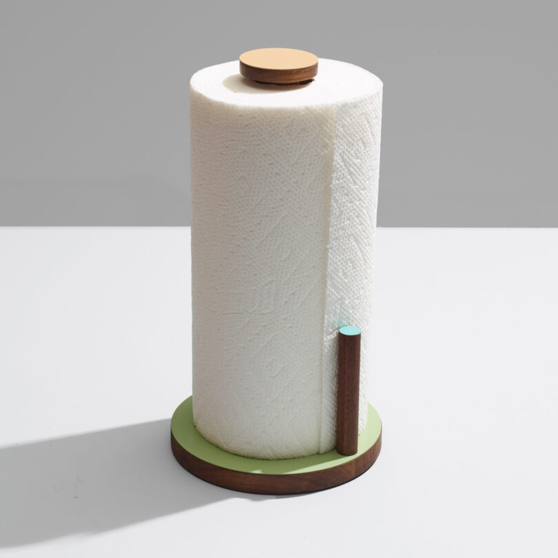 A roll of paper towels on a modern holder with a wooden base and a small vertical rod on a light gray surface