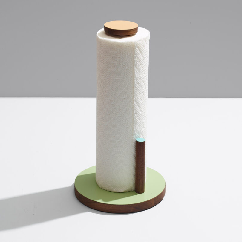 A roll of white paper towels stands upright on a wooden base with a circular holder, placed against a gray background