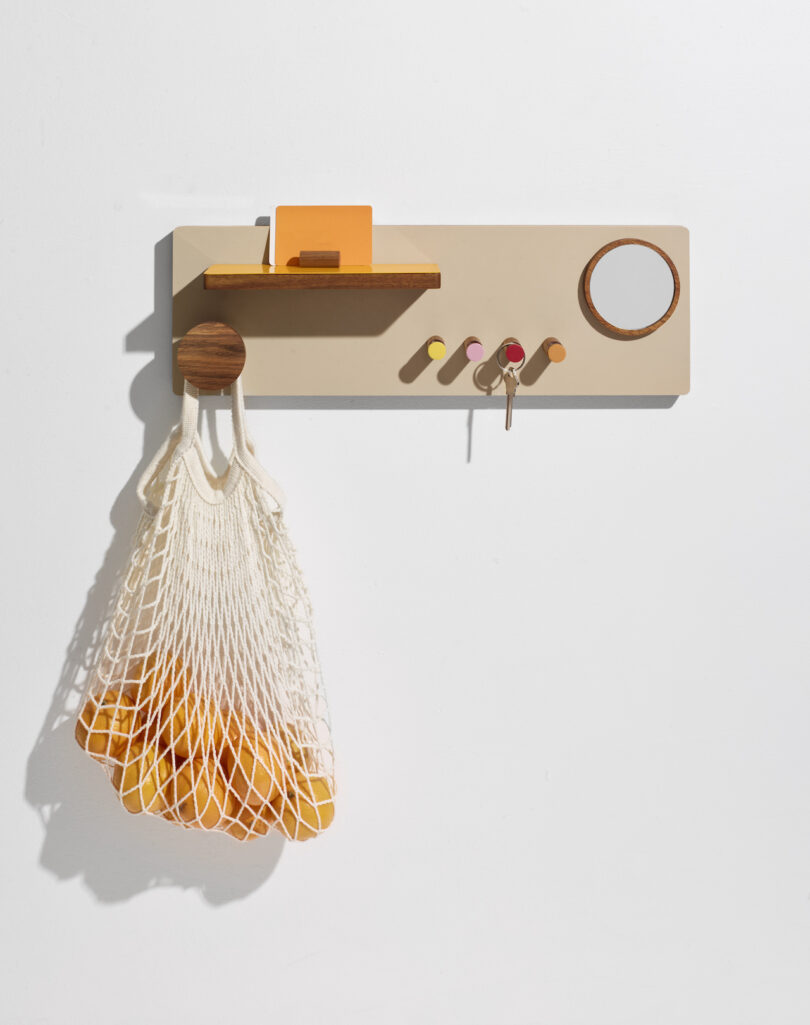 A wall organizer with a small shelf holding a note, hooks with keys, a round mirror, and a hanging net bag containing oranges