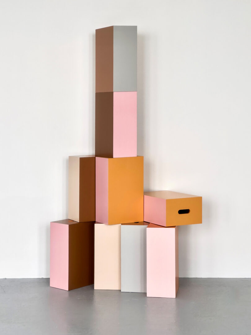 Stacked geometric blocks in shades of pink, orange, gray, and beige against a white wall