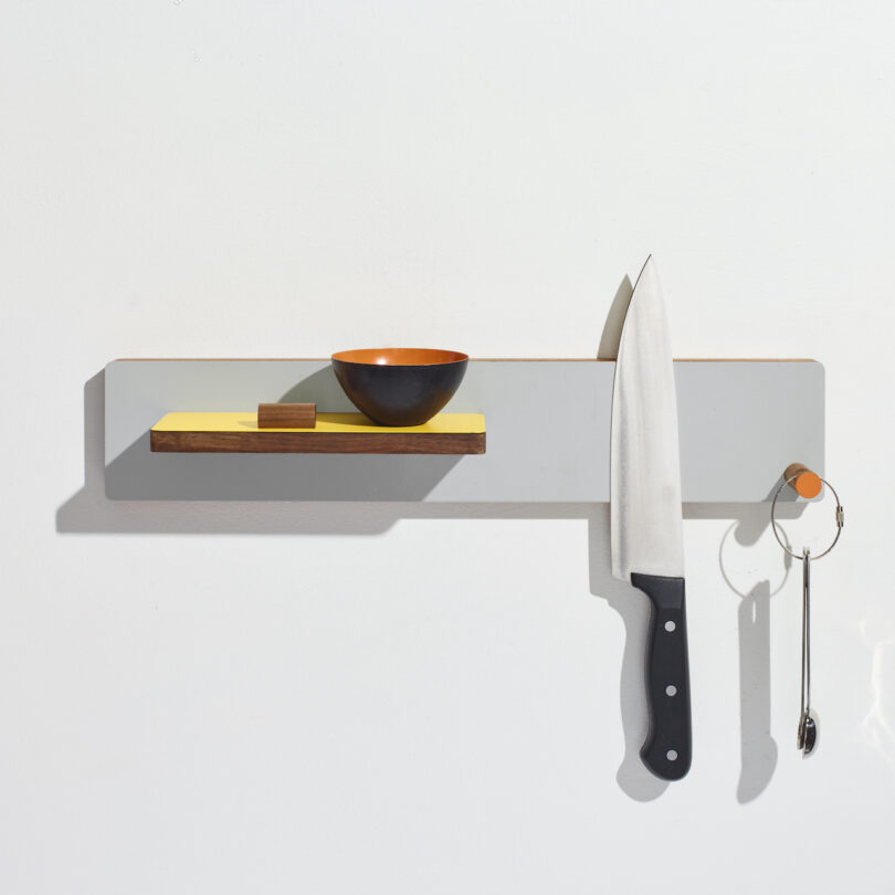 Wall-mounted kitchen organizer with a knife, black bowl, small yellow shelf, and a hanging spoon on a white background