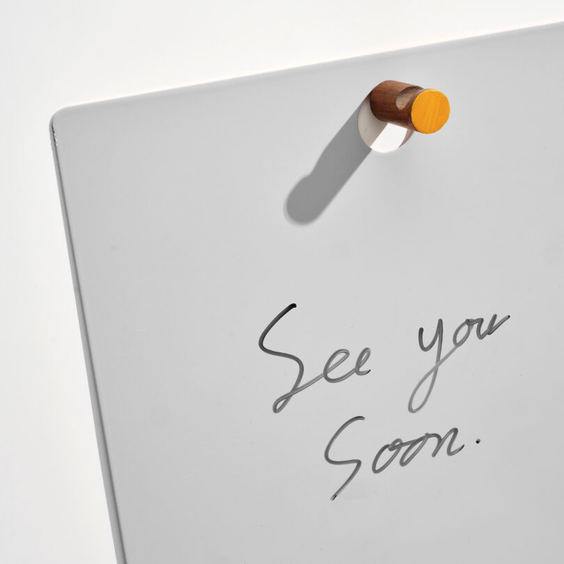 A whiteboard with the handwritten message "See you soon." in black ink, attached to a wall with a round wooden magnet