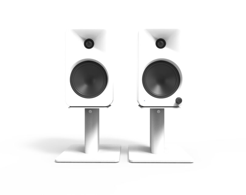 Two white speakers on stands with a minimalist design, each featuring a large woofer and a small tweeter.