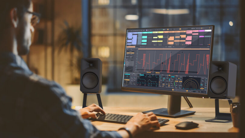 Person working at a computer with audio production software on the screen, surrounded by speakers in a dimly lit room.